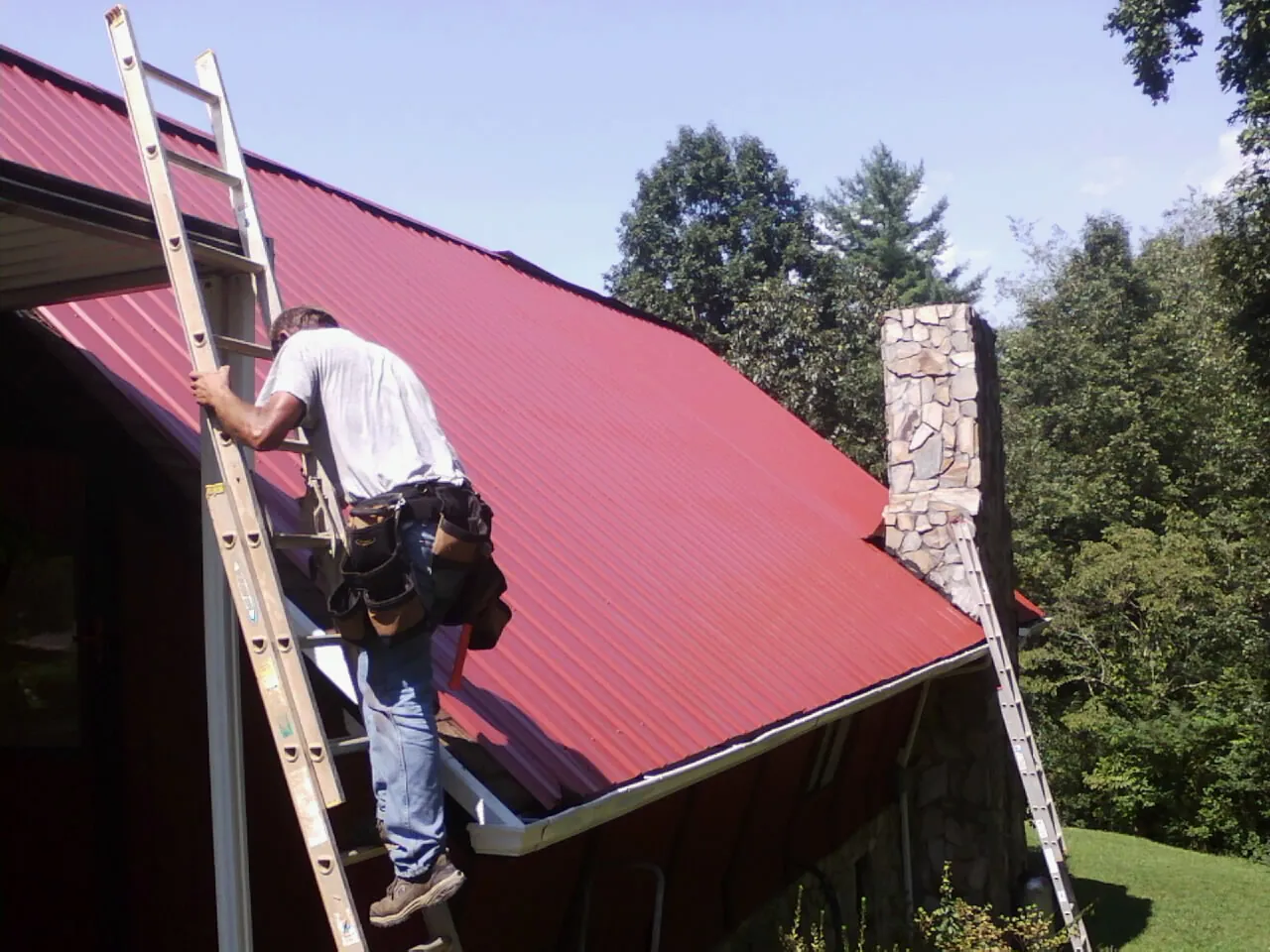 Roofer on a ladder beside a red metal roof and stone chimney — Soffit & Fascia Repair by Roofing Brothers in North Dartmouth